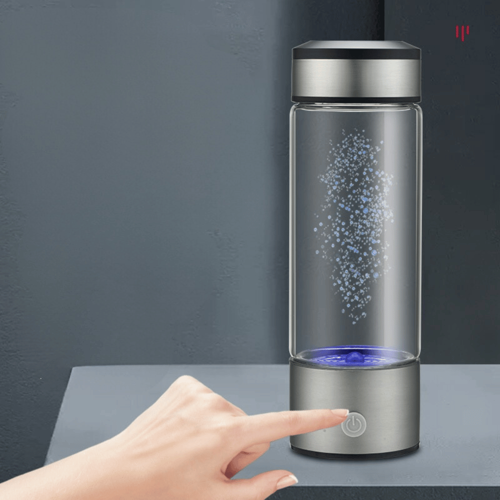 Glass water bottle with silver base and black lid on a dark surface, with a hand touching the button.
